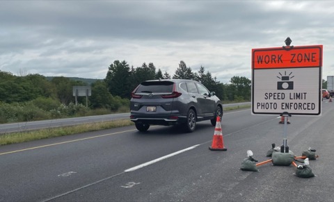 A roadside sign reading WORK ZONE, SPEED LIMIT PHOTO ENFORCED, with a camera symbol.