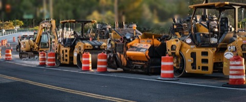 A line of construction equipment such as pavers and rollers staged in a work zone.